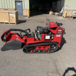 Red Toro snow blower with a clear windshield sits outside a workshop, surrounded by pallets, boxes, and industrial equipment on a sunny day.