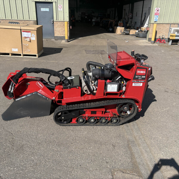 Red Toro snow blower with a clear windshield sits outside a workshop, surrounded by pallets, boxes, and industrial equipment on a sunny day.