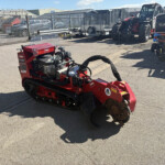 Red Toro stump grinder in a paved outdoor yard with a Kubota tractor on the right and metal fencing in the background.