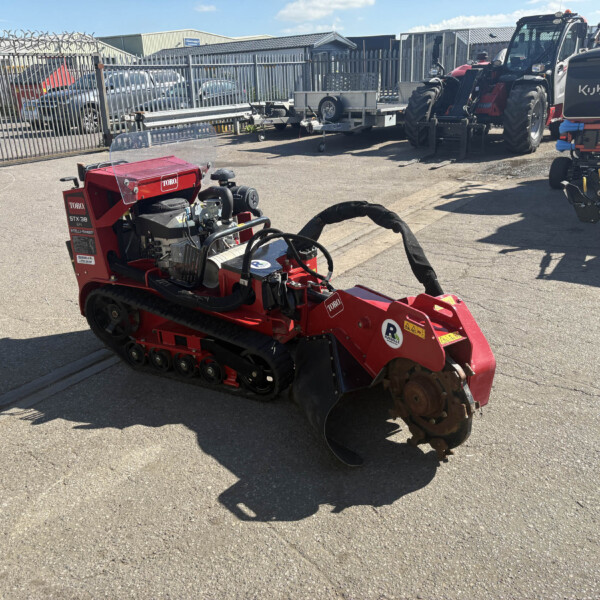 Red Toro stump grinder in a paved outdoor yard with a Kubota tractor on the right and metal fencing in the background.