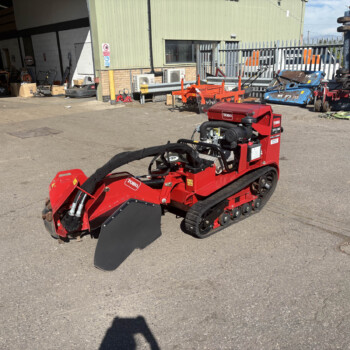 Red Toro tracked machine with a front blade parked in an outdoor equipment yard. Behind it is a green industrial building and various tools and machinery.