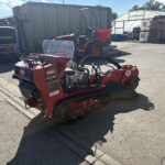 Red Toro STX-38 tracked machine in a sunny industrial yard, with pallets, a greenhouse, and a large metal container nearby.