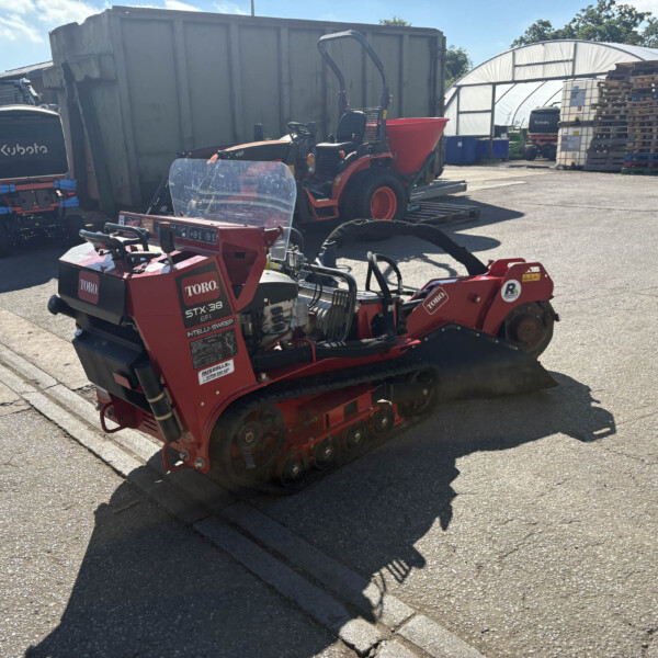 Red Toro STX-38 tracked machine in a sunny industrial yard, with pallets, a greenhouse, and a large metal container nearby.