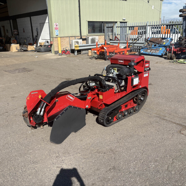 Red Toro tracked machine with a front blade parked in an outdoor equipment yard. Behind it is a green industrial building and various tools and machinery.