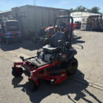 Red Toro ride-on mower on a trailer in a yard with pallets and industrial equipment nearby.