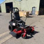 Ride-on Toro mower with red mowing attachment in a parking lot beside a large corrugated metal warehouse and open loading bay.