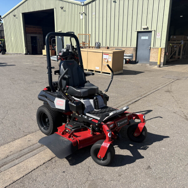 Ride-on Toro mower with red mowing attachment in a parking lot beside a large corrugated metal warehouse and open loading bay.