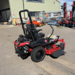 Red and black ride-on lawn mower parked on asphalt in an industrial yard with a green metal building and scattered equipment in the background.