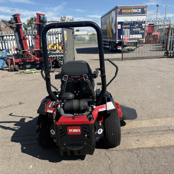 Front view of a red Toro ride-on mower in a paved yard, with industrial equipment and a tall metal fence in the background.