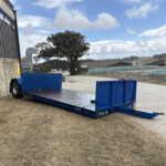 Blue flatbed trailer in an industrial yard beside a brick wall and grey warehouses under a partly cloudy sky