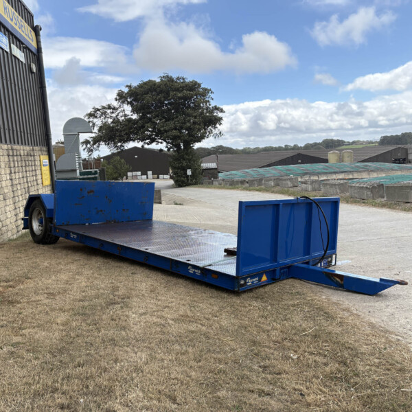 Blue flatbed trailer in an industrial yard beside a brick wall and grey warehouses under a partly cloudy sky