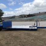 Blue flatbed trailer on dry grass at an industrial yard with buildings and tarped ground in the distance under a cloudy sky.