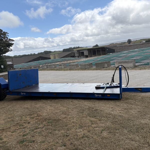 Blue flatbed trailer on dry grass at an industrial yard with buildings and tarped ground in the distance under a cloudy sky.
