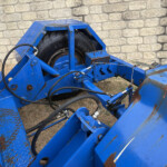 Blue industrial machinery with a large black tire, rust, and hydraulic hoses visible against a stone wall backdrop.