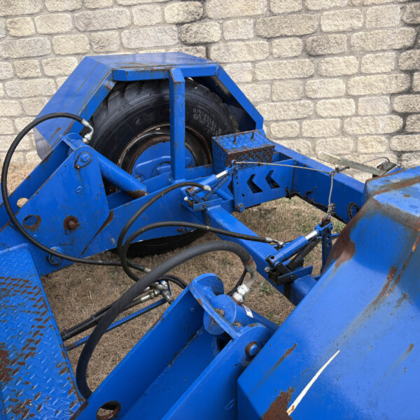 Blue metal agricultural/industrial machine frame with a large black tire, hydraulic hoses, and rust spots, set against a stone wall.