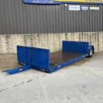 Blue flatbed trailer parked in front of a grey corrugated metal building with a brick lower wall and signs on the wall.