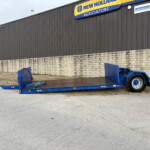 Blue flatbed trailer parked on a concrete lot in front of a gray corrugated building with a New Holland Agriculture sign at the top edge of the wall.