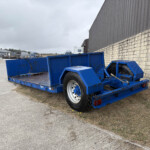 Blue flatbed trailer in the foreground near a stone wall on a cloudy day at an industrial yard.