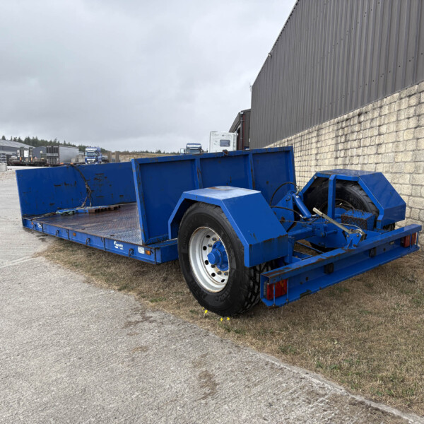Blue flatbed trailer in the foreground near a stone wall on a cloudy day at an industrial yard.
