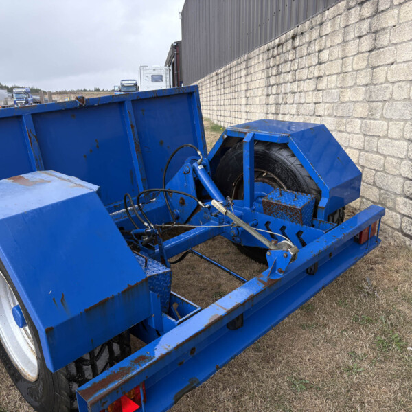 Blue industrial trailer with rust, parked beside a stone wall in a yard, distant trucks in the background.