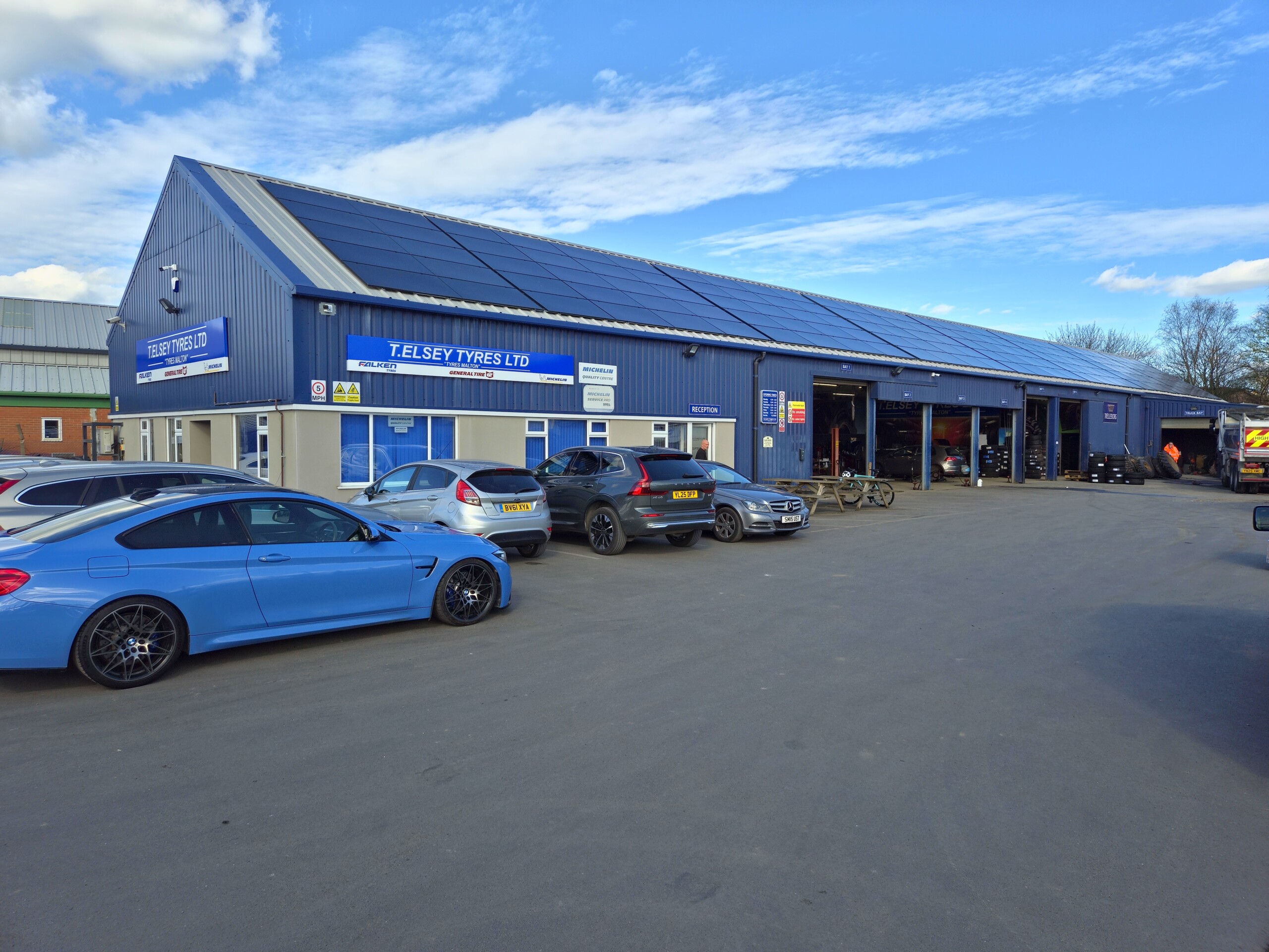 Blue industrial building with solar panels on the roof, a row of blue cars parked in front, and a sign for T. Elsey Tyres Ltd in view.