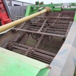 Green agricultural conveyor/header with rusted metal tines and a beige PTO drive shaft extending from a red tractor on the left; weathered and dusty equipment outdoors.
