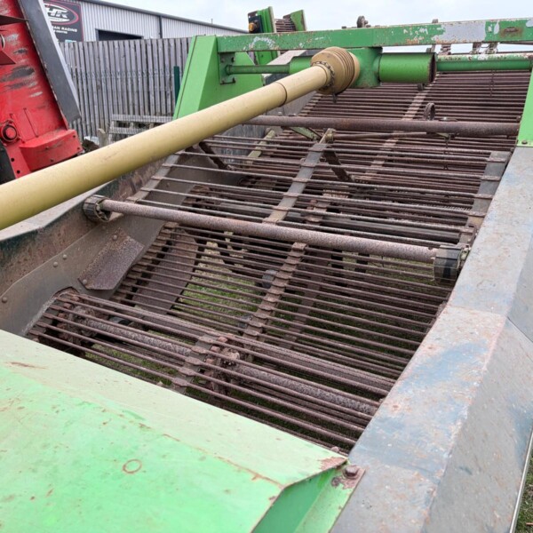 Green agricultural conveyor/header with rusted metal tines and a beige PTO drive shaft extending from a red tractor on the left; weathered and dusty equipment outdoors.