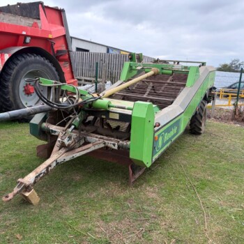 Green farming implement attached to a red dump truck, with a PTO drive shaft and hydraulic hoses, resting on grass near a fence.