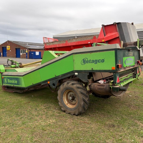 Green Netagco/Reekie agricultural machine with a red hopper parked on a grassy area near a workshop and van in the background
