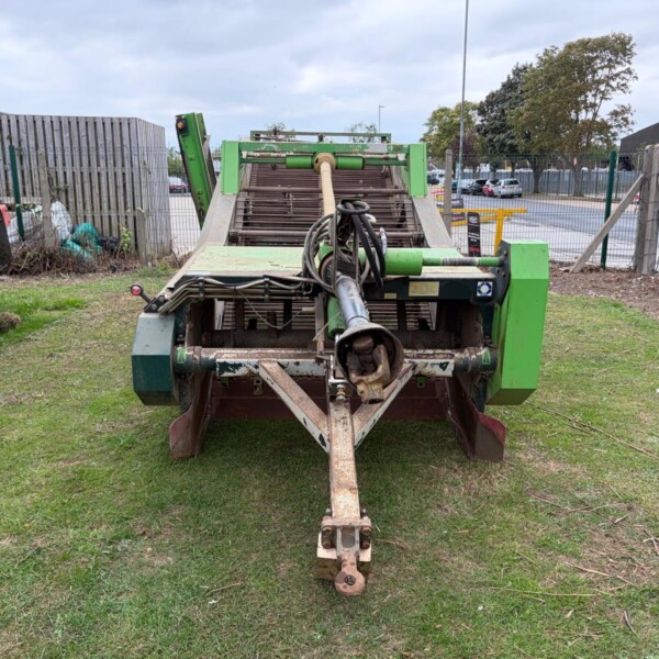 Tow-behind green farm implement with roller conveyor and hitch, parked on grass beside a fence and truck tire. Remember to include power shaft and hoses.