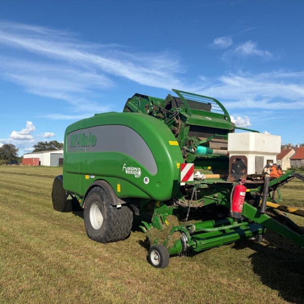 Green McHale Fusion baler attached to a tractor on a grassy farm field under a blue sky with scattered clouds.