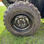 Close-up of a large knobby mud-terrain tire on a black wheel, attached to an off-road vehicle, with grass in the background.