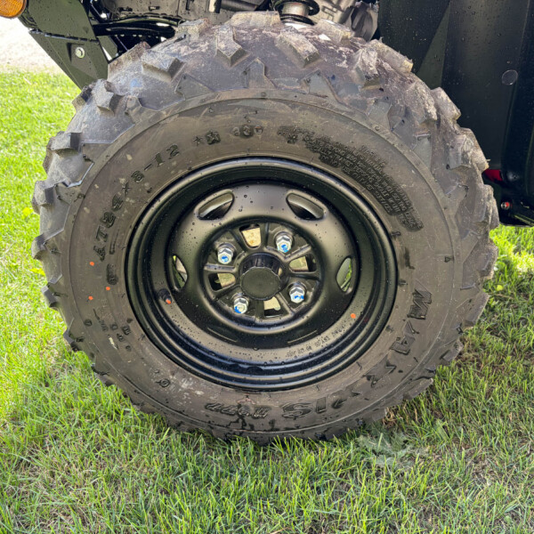 Close-up of a large knobby mud-terrain tire on a black wheel, attached to an off-road vehicle, with grass in the background.