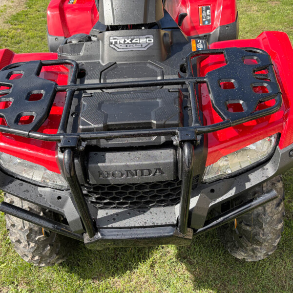 Red Honda ATV front with a black cargo rack, headlights visible, on grassy ground; wet with water droplets.