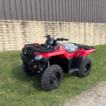 Red all-terrain vehicle (ATV) parked on grass beside a stone wall and brown metal building exterior. wide-angle outdoor scene.