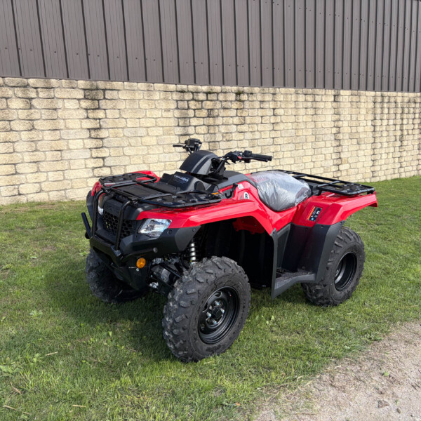 Red all-terrain vehicle (ATV) parked on grass beside a stone wall and brown metal building exterior. wide-angle outdoor scene.