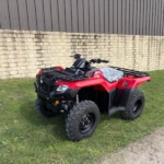 Red ATV parked on grass beside a stone wall and a gray metal building, with a yellow sign and an orange cone in the background.