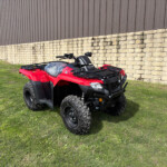 Red ATV parked on a grassy area beside a beige stone wall and brown metal building in the background.