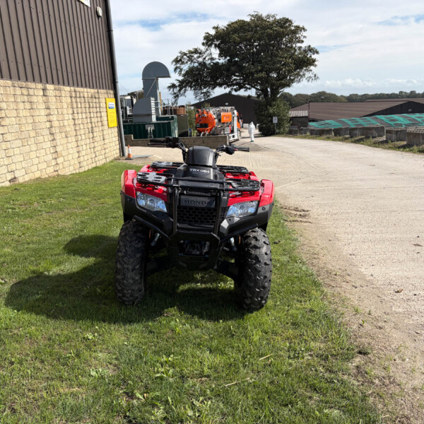 Red Honda ATV facing camera parked on a grassy verge beside an industrial building and stone wall; distant worker in white coveralls near the road.