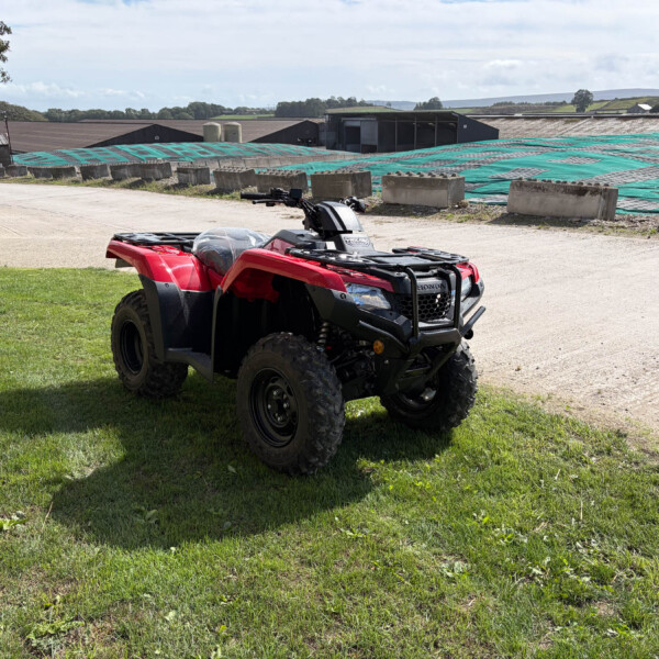 Red ATV on a grassy patch with industrial buildings and tarped ground in the background; a person in white protective clothing stands left.