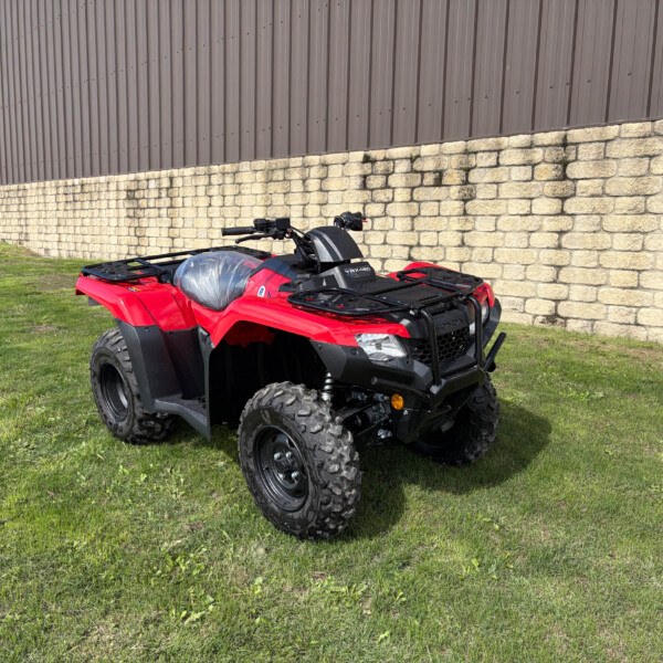 Red ATV parked on a grassy patch beside a tall stone wall and a brown metal building in the background.