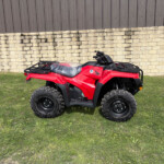 Red Honda ATV parked on grass beside a beige stone wall and brown metal fence, outdoor daytime scene.