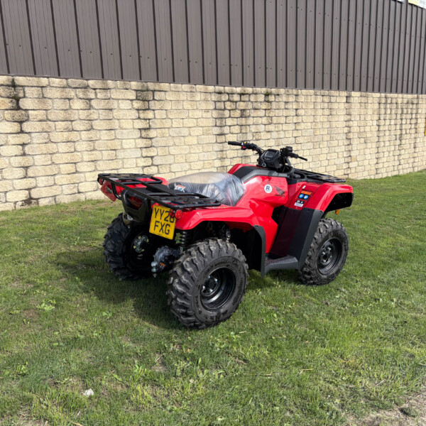 Red all-terrain vehicle parked on grass beside a stone wall and brown metal building, facing left.