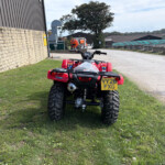 Rear view of a red ATV/quad bike parked on grass by a brick wall and industrial buildings, license plate YY26 FGX visible.