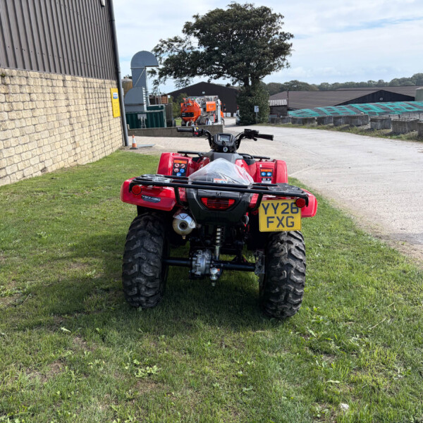 Rear view of a red ATV/quad bike parked on grass by a brick wall and industrial buildings, license plate YY26 FGX visible.