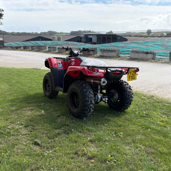Red ATV parked on a grassy field with industrial buildings and tarped yards in the background.