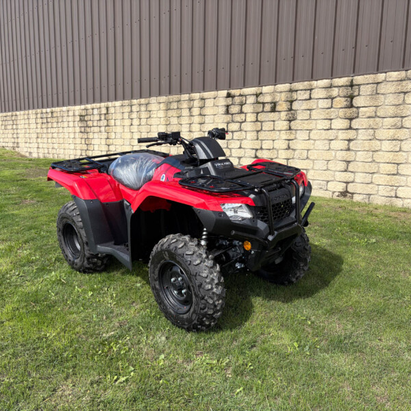 Red ATV parked on a grassy area beside a beige stone wall and brown metal building in the background.