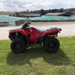 Red Honda ATV parked on a grassy field with farm buildings and gravel driveway in the background.