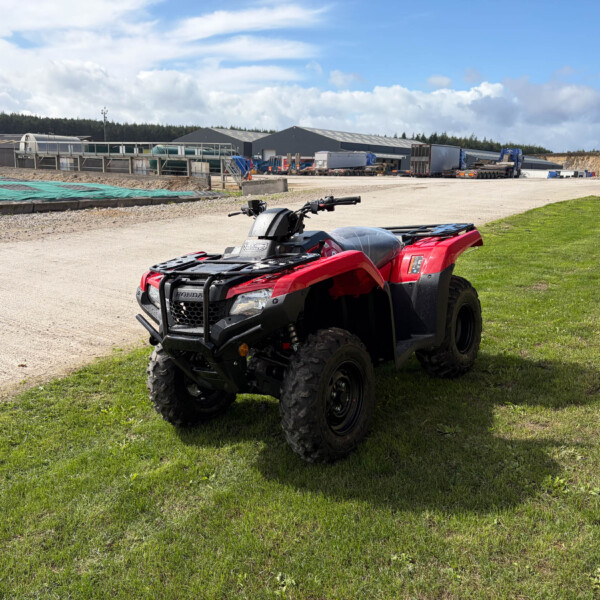 Red ATV parked on a grassy field with an industrial yard and trucks visible in the background under a blue sky.​