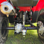 Underside of a red off-road vehicle showing large tires, suspension, and exhaust on grass.